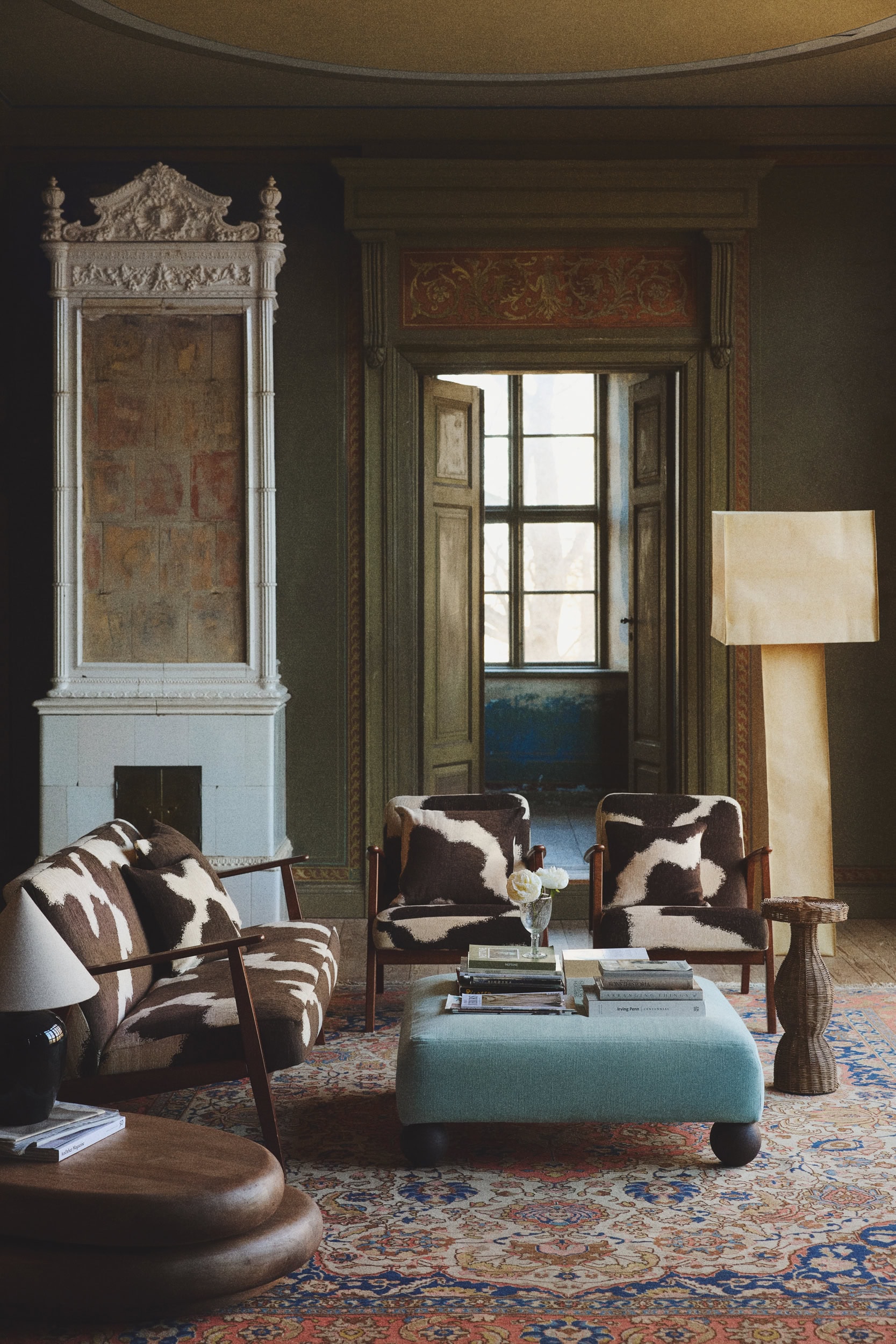 Image of a brown and white, cow print-looking sofa with a traditional red and blue rug underneath and a blue coffee table with stacks on magazines on it.