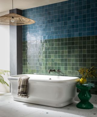 Bathroom with blue and green gloss tiled wall, large white bathtub, bright green stool, vase with flowers, large, naturally textured pendant, white and gray terrazzo tile flooring