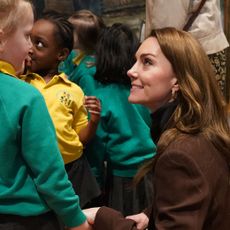 Kate Middleton wearing a brown blazer bending down to smile at a group of school kids in teal sweatshirts