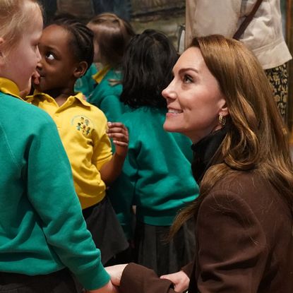 Kate Middleton wearing a brown blazer bending down to smile at a group of school kids in teal sweatshirts