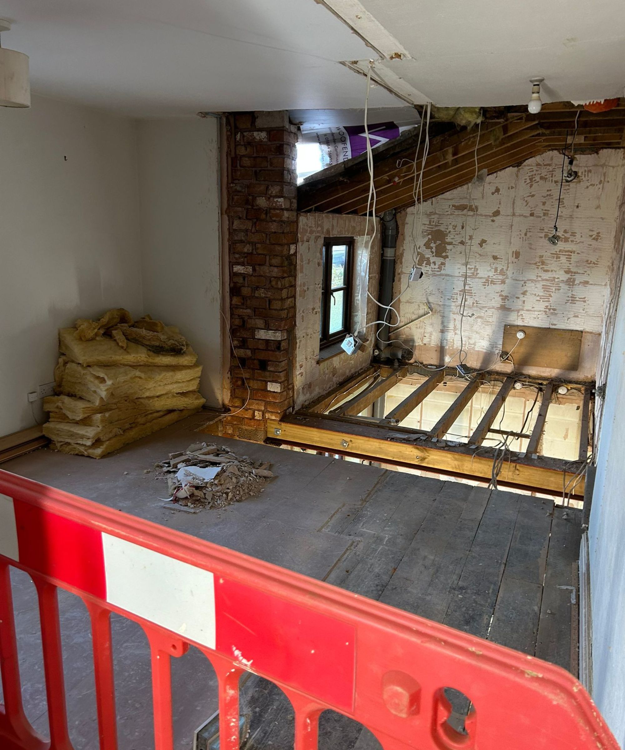 An upper-floor room undergoing renovation, with exposed floor joists, stripped walls and loose wiring hanging from the ceiling. A red safety barrier separates the walkway from the open joists, while insulation and debris sit stacked to one side.