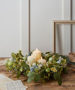 A green leafy wreath with daisies and blue flowers on it and three candles in the middle of it, on a dark brown wooden table with two pieces of paper next to it, in front of a white wall with panelling