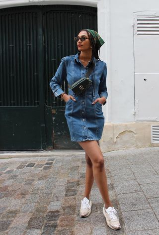 A woman's jean dress outfit with a button-down dress with a scarf, sunglasses, a black crossbody bag, and white sneakers.