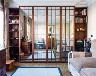 Family room space with striking paneled mirror wall, dark wooden panels and shelving units filled with books, dark wood parquet flooring, black and cream rug, beige sofa, dark wood desk and storage units