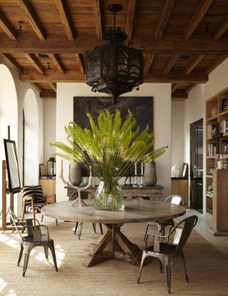 High exposed wooden ceiling, cream walls with large window arches, large circular table with large plant in the centre and rustic decorations