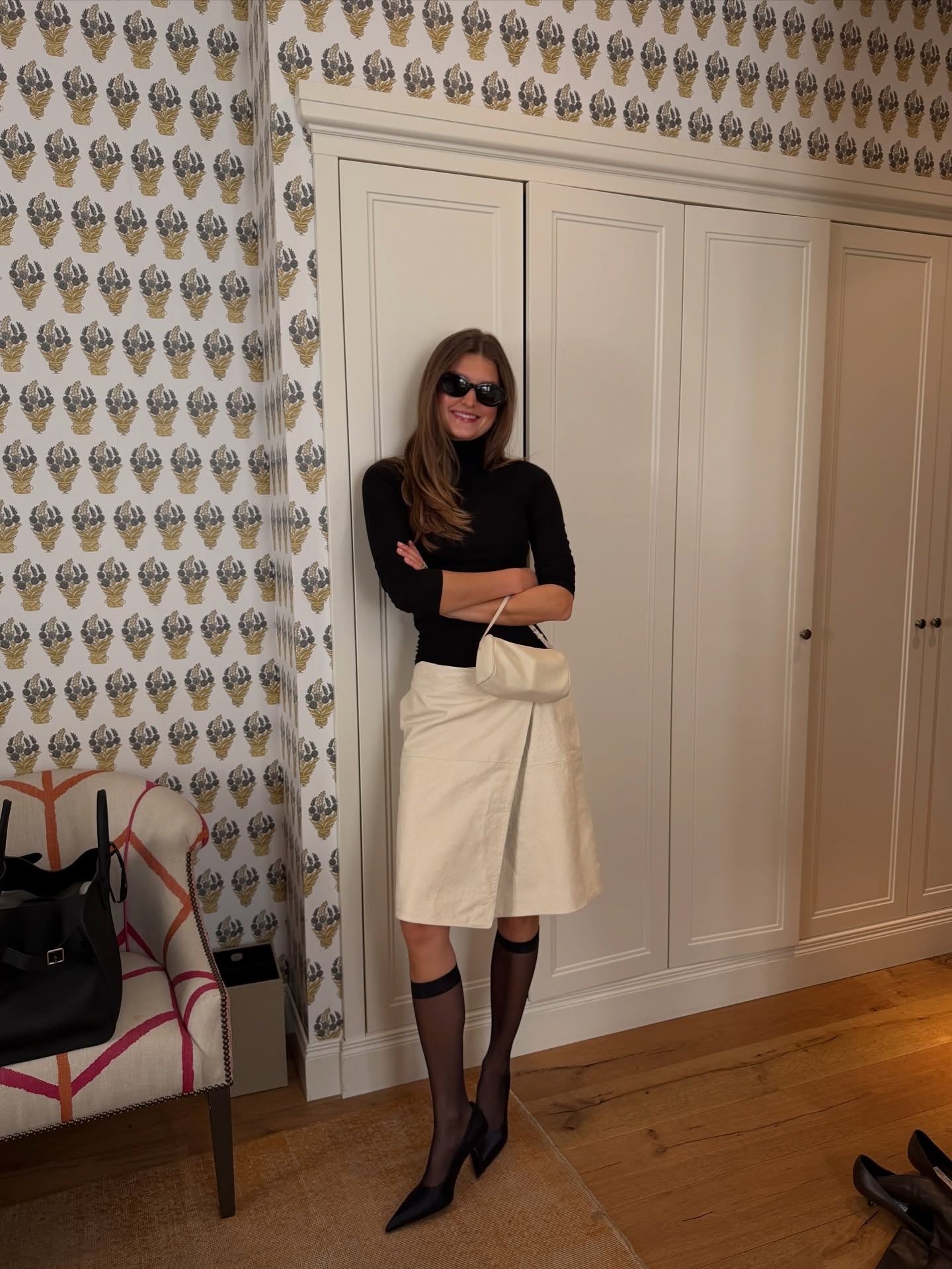 Fashion person Sara Walker wears a transitional winter to spring outfits 2026. She wears a black thin knit roll neck, cream midi skirt, black stockings and black pointed toe heels. She accessories with oversized sunnies and The Row '90s bag. She is stood in front of a white closed wardrobe in a recent image from her Instagram.
