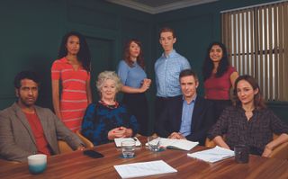 The cast of Ten Percent: seated around a table from left to right are Prasanna Puwanarajah as Dan, Maggie Steed as Stella, Jack Davenport as Jonathan and Lydia Leonard as Rebecca. Standing behind them from left to right are Fola Evans-Akingbola as Zoe, Rebecca Humphries as Julia, Harry Trevaldwyn as Ollie and Hiftu Quasem as Misha. 