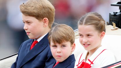 Prince George and Prince Louis wearing blue suits and red ties and Princess Charlotte wearing a red-and-white dress with a red bow riding in a carriage