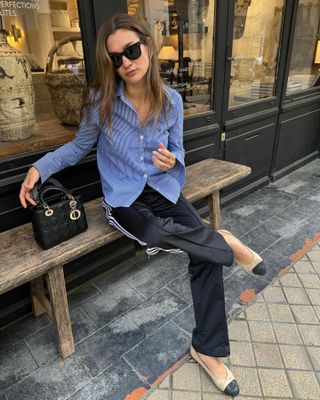 The French style influencer sat on a bench outside a Paris cafe in a blue striped button-down shirt, Dior bag, Adidas sweatpants and Chanel cap-toe ballet flats