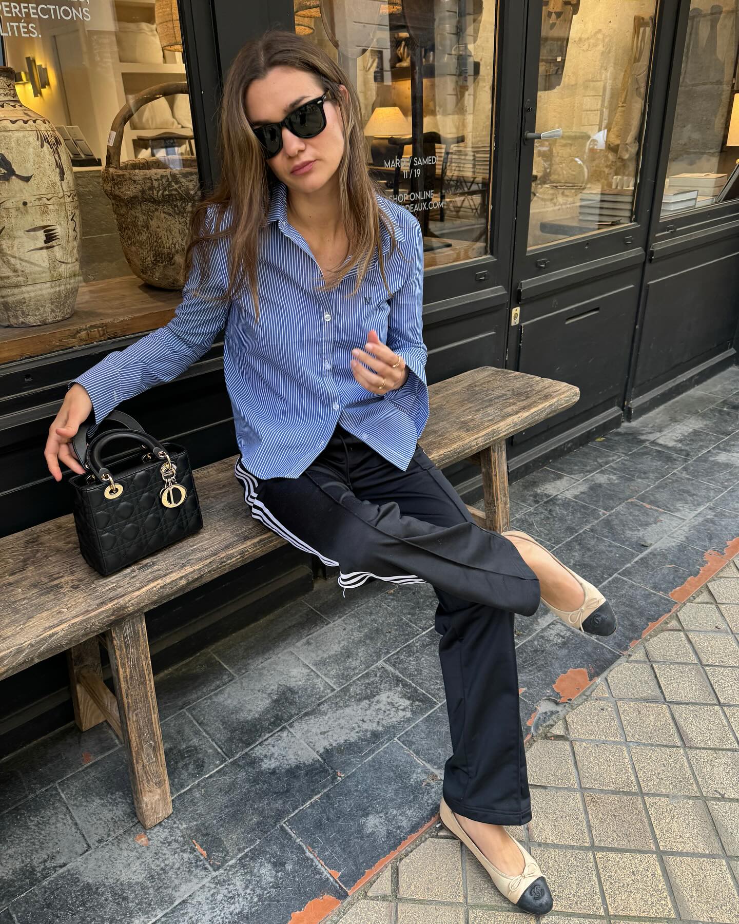 French style influencer sits on a bench outside of a Parisian cafe in a striped blue button-down shirt, Dior bag, Adidas track pants, and Chanel cap-toe ballet flats