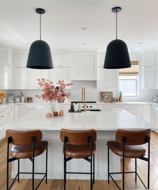 A white kitchen with white cabinets, a marble kitchen island with faucet, soap dispensers and a vase with leaves in it, three leather bar stools and two pendant lights hanging from the ceiling