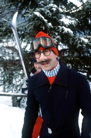 King Charles wearing a black ski suit walking through the snow with fake glasses, a fake mustache and comedy nose