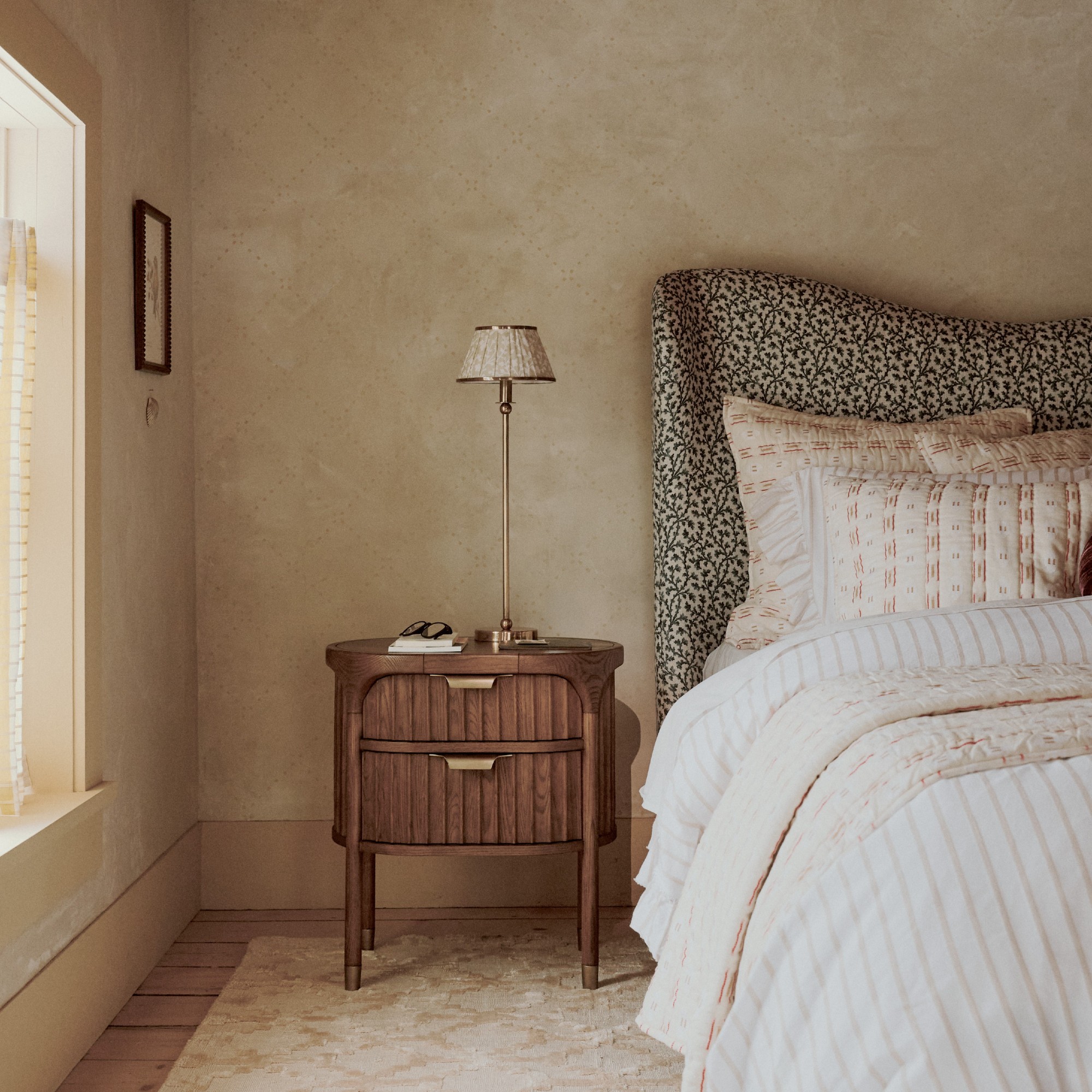 A limewash-painted bedroom with a patterned upholstered curved headboard and a wireless table lamp on the bedside table