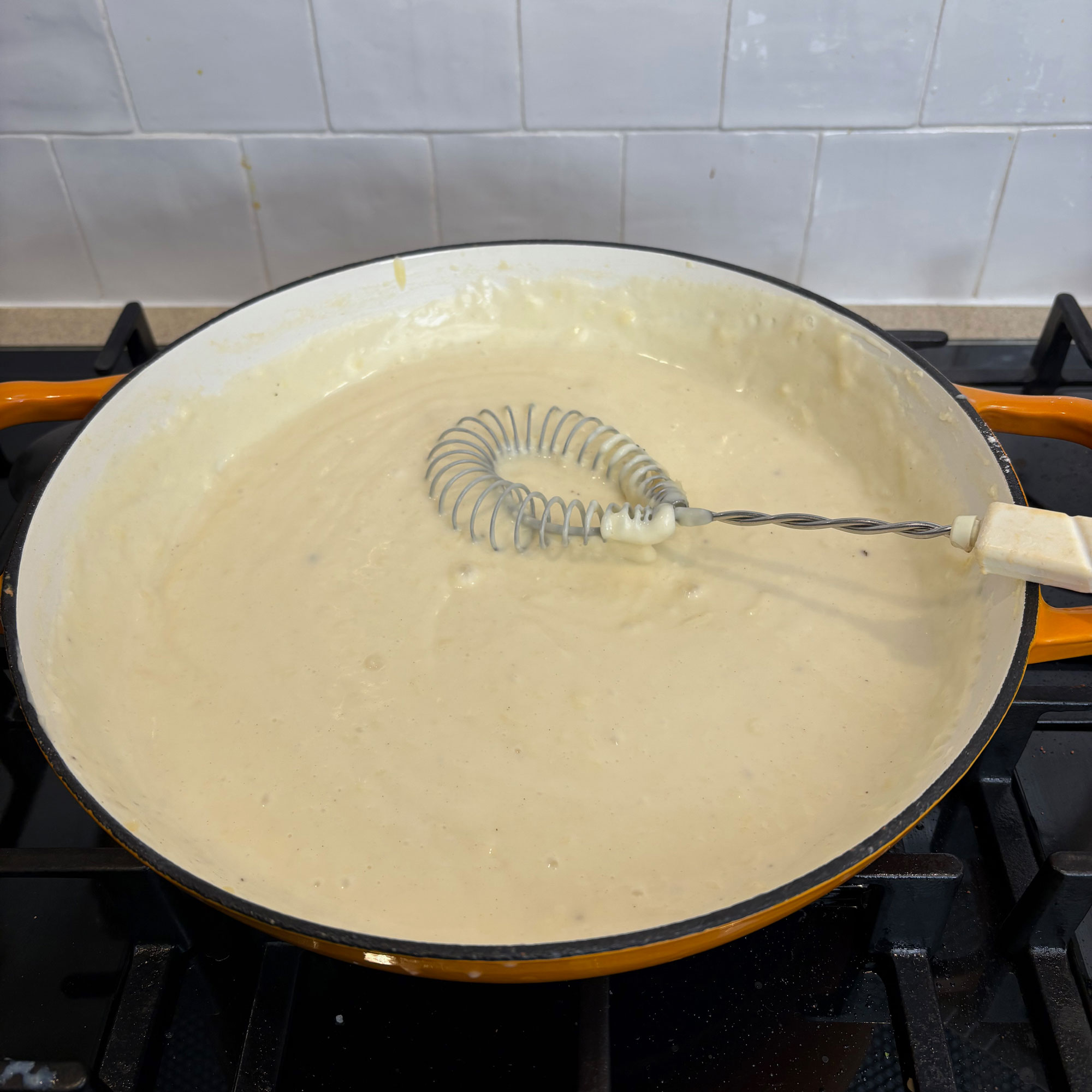 Orange cast iron pan on black hob with cheese sauce and metal whisk