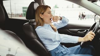 A woman with light brown hair and wearing a blue shirt yawns while driving her car