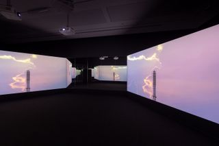 A video installation. The screens show a bright, pink sky with a top part of a metal tower.