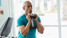 A man in a t-shirt holds a dumbbell to his chest. Behind him we see the rear of a screen attached to an exercise bike.