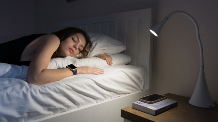 A female student sleeping in bed wearing wrist sleep tracker next to bedside table with lamp, book and phone