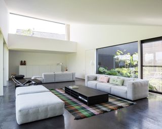 A lounge area with floor to ceiling windows and doors, white walls, white ceilings and vinyl floors with a tartan rug set beneath a black coffee table in between a grey three-sater sofa and 2 large cushions. on the far end is a grey three-seater sofa , facing the white cabinet resting against a white wall.