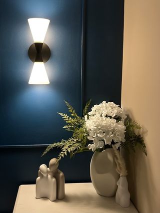 a side table decorated with flowers and ornaments next to a dark blue feature wall with a lit sconce