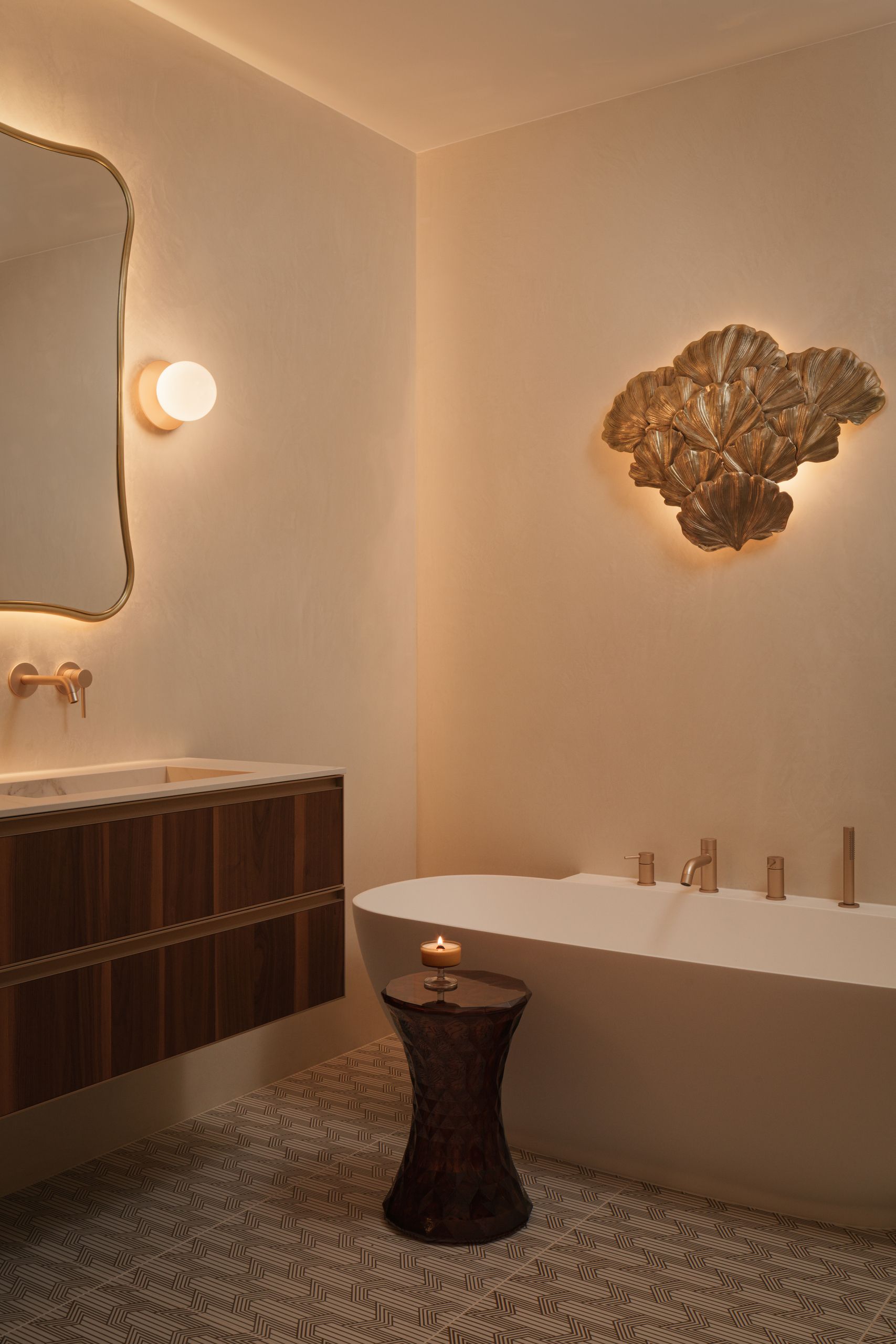 Image of a luxurious, calming, and modern bathroom. A big white bathtub, gold hardware, a gold, sculptural sconce, a side table with a candle on it.