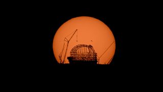 A large orange sun illuminates the scaffolding of a dome where a telescope is being built, along with cranes next to it.
