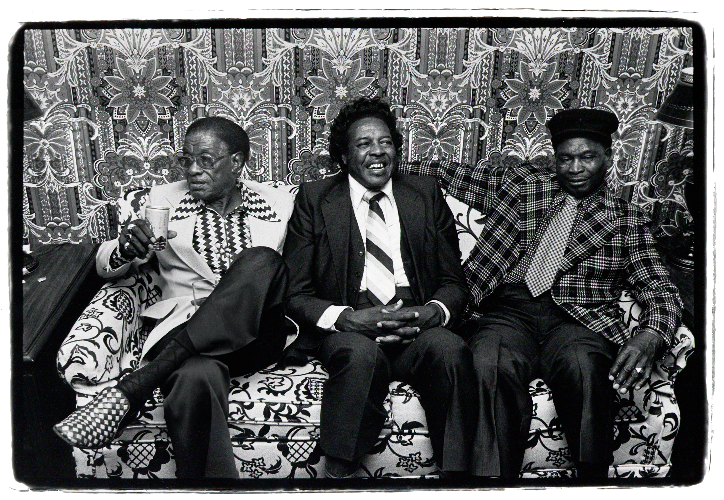 Three older blues musicians in patterned suits sit side by side on a floral sofa in front of ornate patterned wallpaper