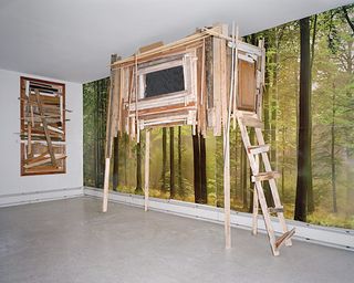 ’Mr. Schuhlmann or the Man in the High Castle’ by Matthieu Lavanchy, 2009. A wooden structure on stilts in front of a wall with a forest photograph on it.
