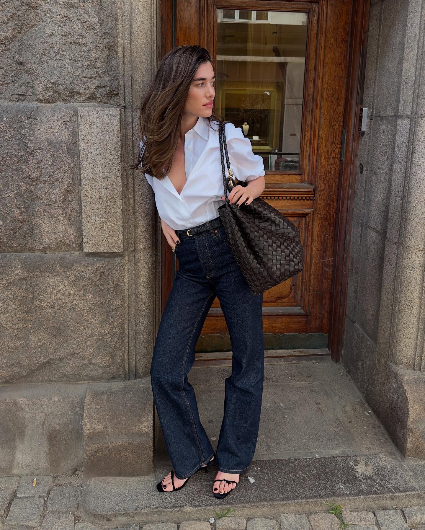 @iliridakrasniqi wearing a white button-down shirt with high-waisted jeans and strappy sandals.