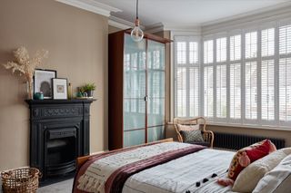 Neutral peach bedroom with wood and glass wardrobe, boho bedding and bay window with white shutters