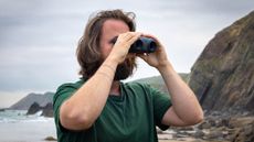 A male holding the Canon 8x20 IS binoculars with a view of the sea and coast behind.