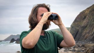 A male holding the Canon 8x20 IS binoculars with a view of the sea and coast behind.