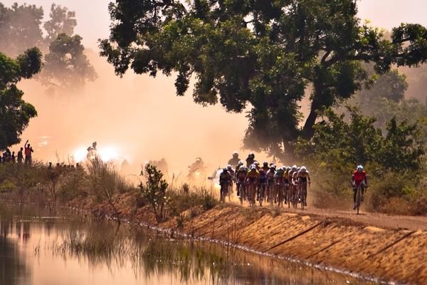 The peloton emerges onto a bridge in a cloud of dust.