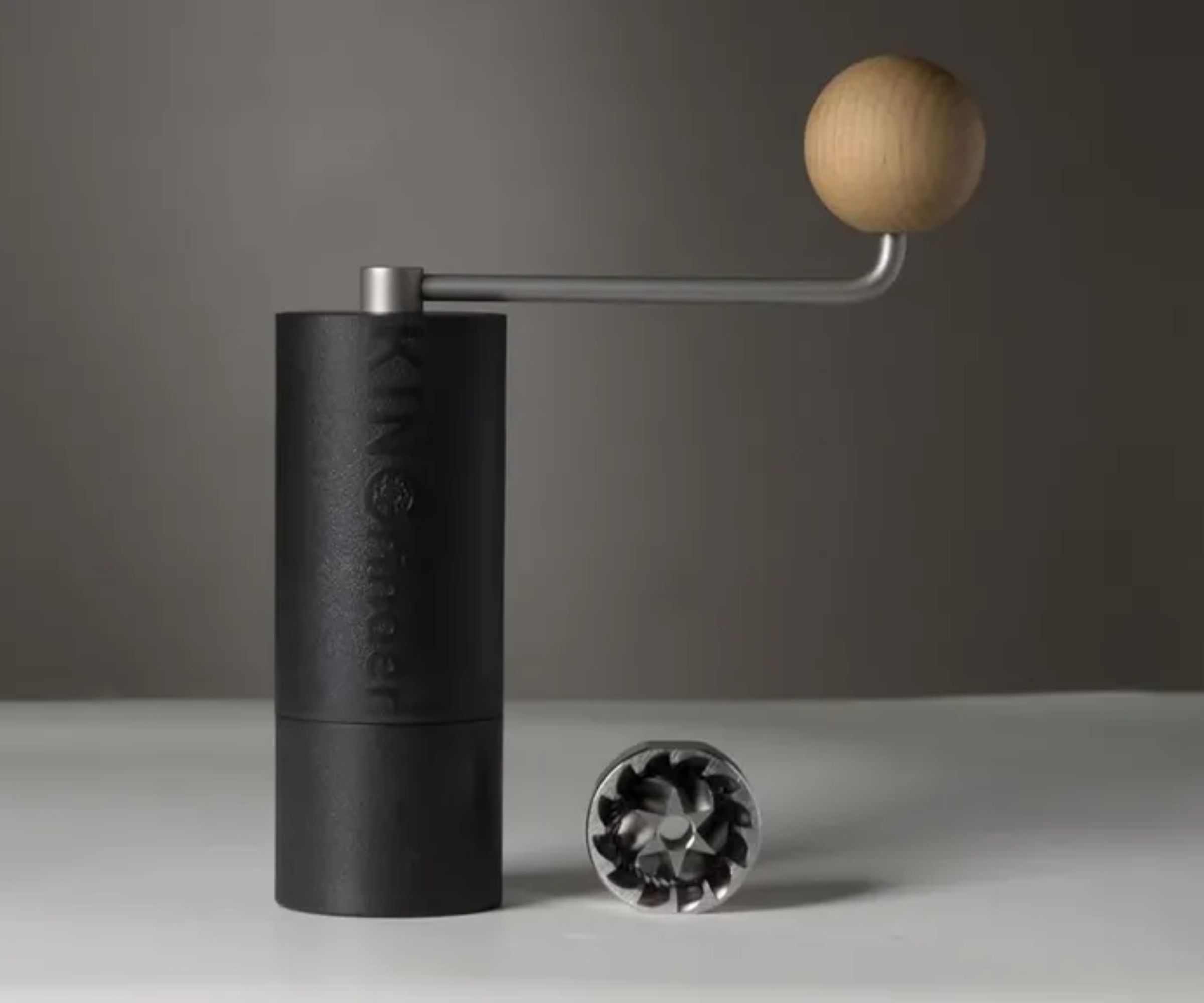 A black cylindrical coffee grinder on a white countertop, with a stainless steel crank handle. 