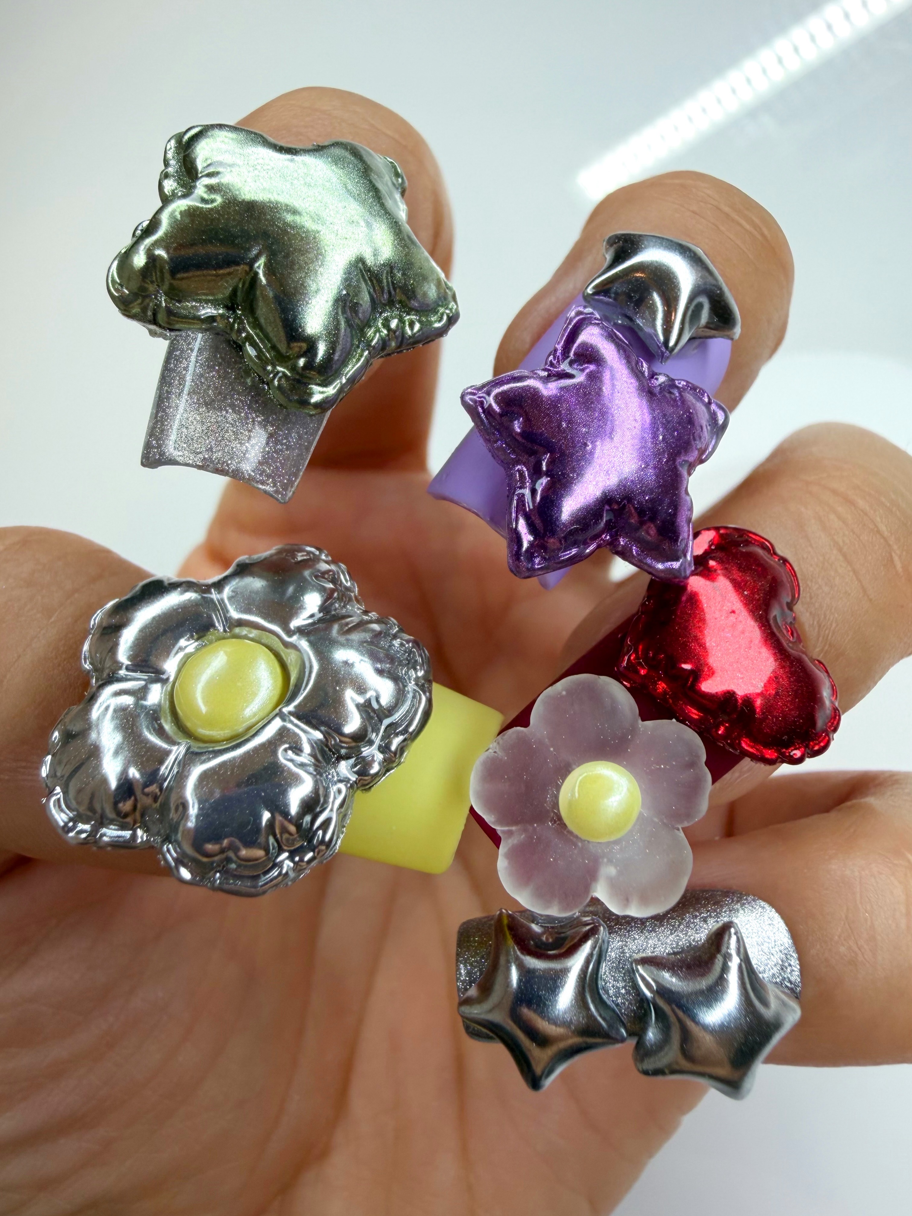 a maximalist manicure with large balloon flowers in silver and yellow, a red heart, and a purple star and green star on a woman's hand with a white background