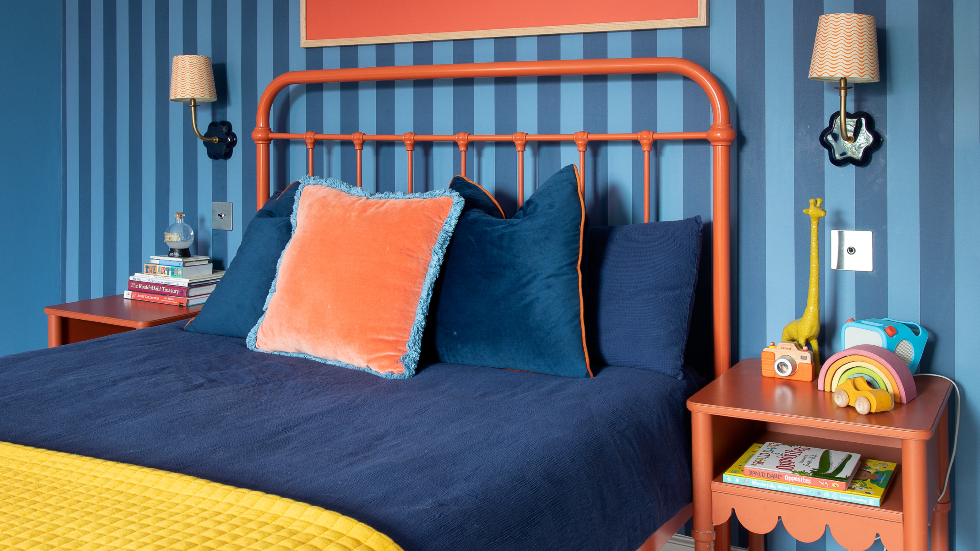 A blue bedroom with a feature wall covered in striped wallpaper paired with a cotal orange metal bedframe and matching bedside tables