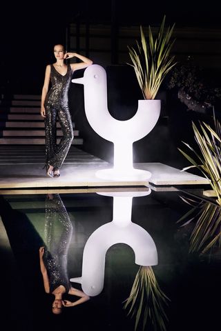 A female model in a black sequin jumpsuit posing bu ta chicken-like sculpture next to a pooll, photographed at night