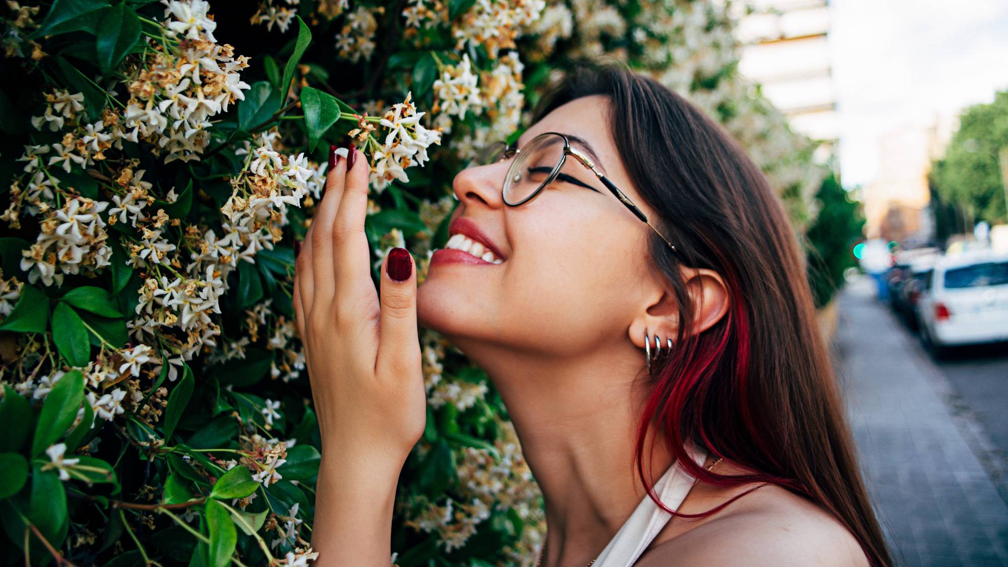 A smiling woman smells a wall of jasmine flowers on a sidewalk