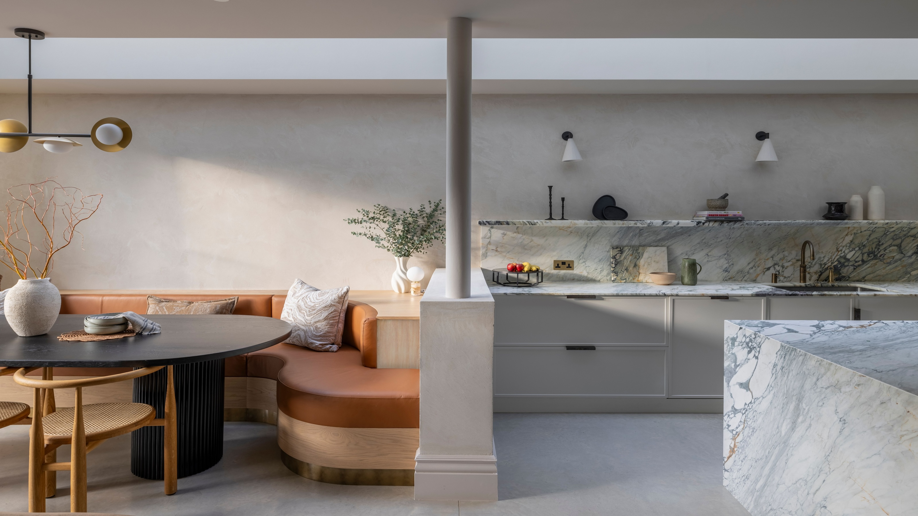 An open plan neutral kitchen with curved banquette seating on one side and a half wall divider with a pillar in the center separating it from the kitchen space on the other side with a large marble kitchen island