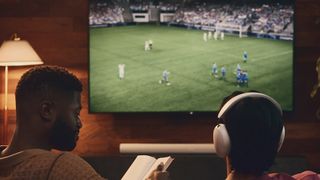 Sonos Ace-hörlurar som bärs av en man som tittar på TV, medan en annan man läser bredvid honom.