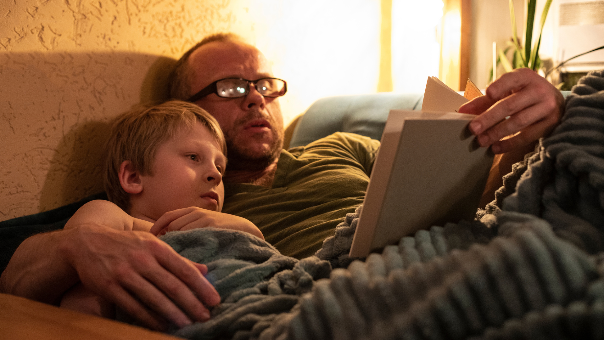 A father and his young son read a book together at night while sitting under a gray blanket.