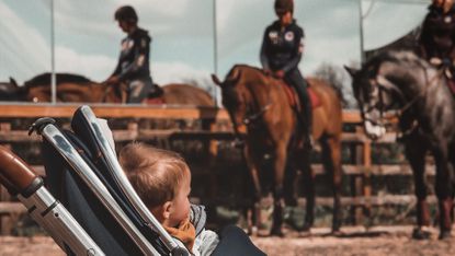 Baby in pram in foreground, with two people riding horses in background after giving birth