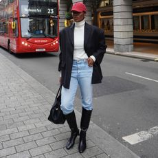 A woman wears a red baseball cap, black blazer, white turtleneck, blue jeans, black knee-high boots, and a black tote bag.