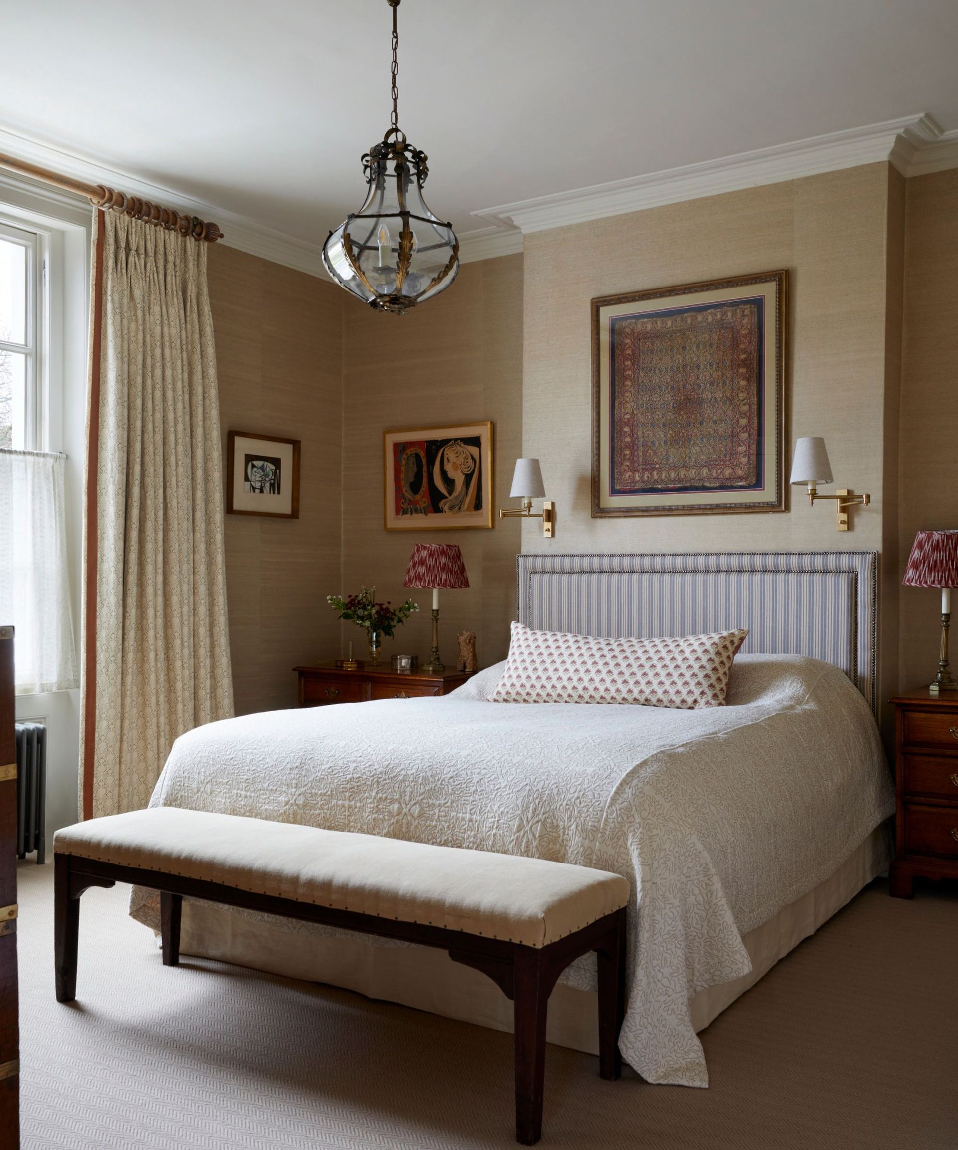 Bedroom with beige walls, low antique pendant, striped headboard, tapestry, sconces and bench