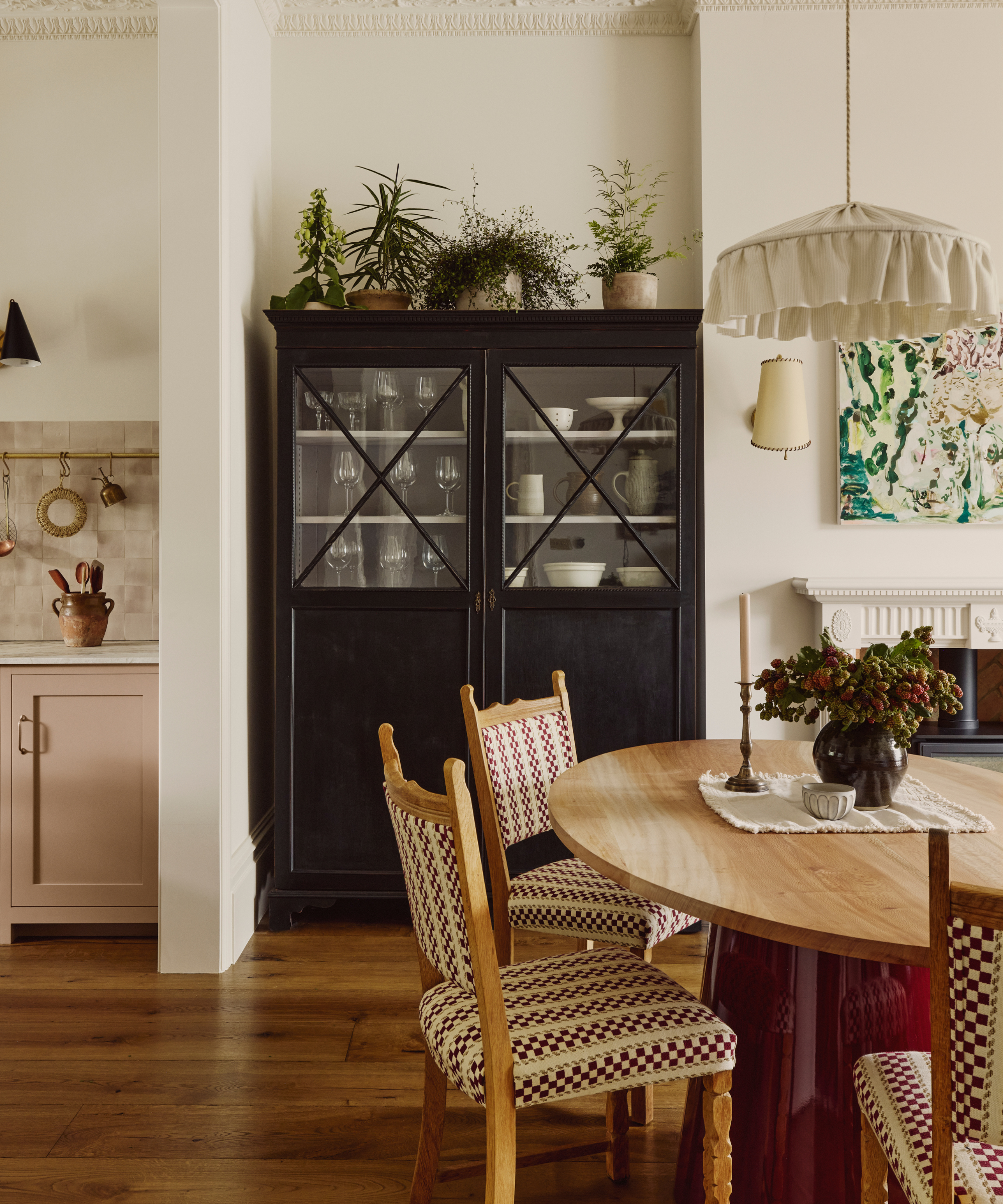 a dining area with a large black cabinet with glassware inside, a wood table with chairs that have patterned upholstery, a fabric pendant hanging from the ceiling, and plants on the cabinet and table