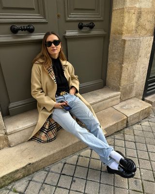 French woman wears a Burberry trench coat, black sweater, jeans and loafers with white crew socks.