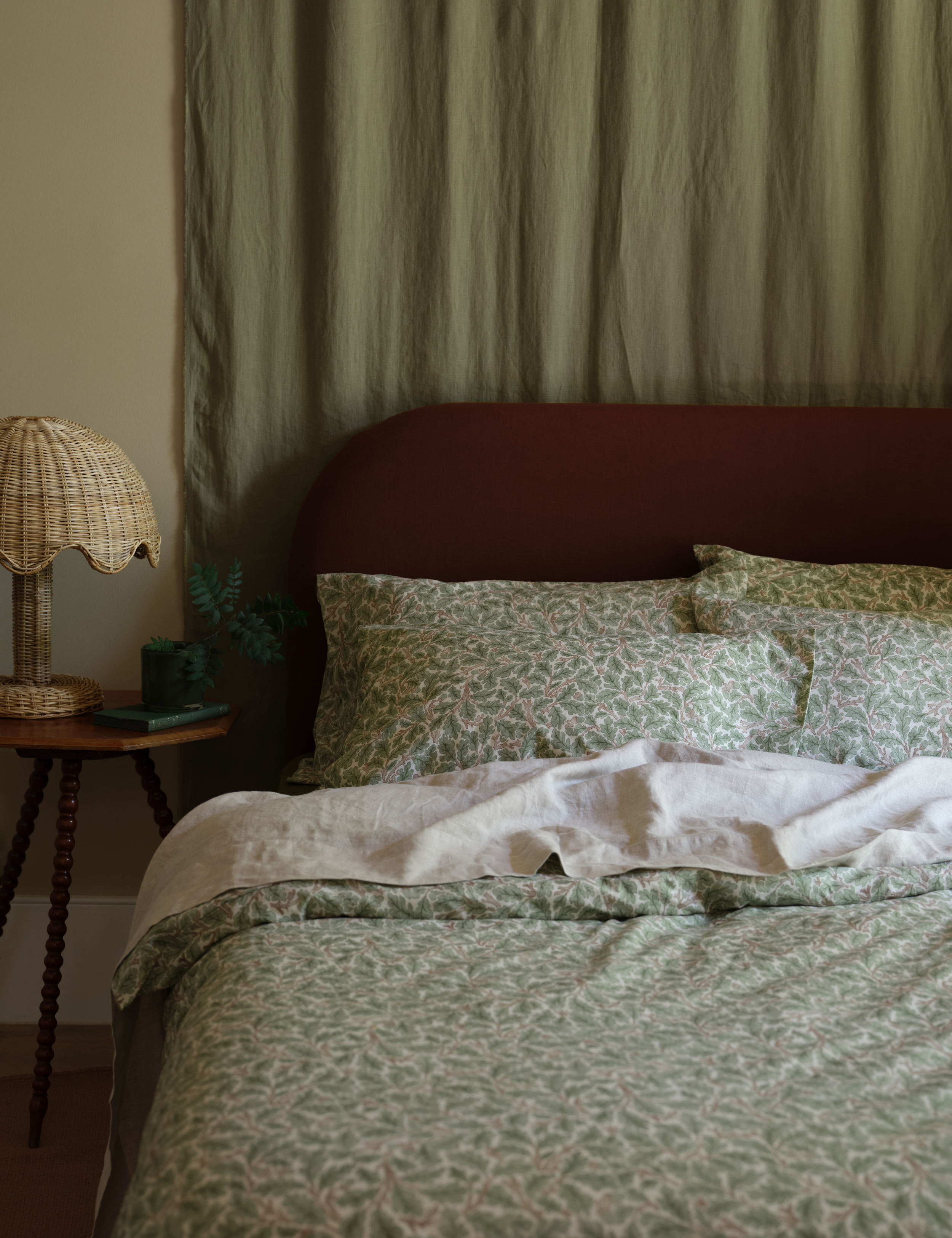 Green botanically patterned Piglet in Bed x Morris &amp;amp; Co. bedding styled in a neutral bedroom with similarly toned wall curtains, a wooden bobbin-legged nightstand, and woven scalloped table lamp
