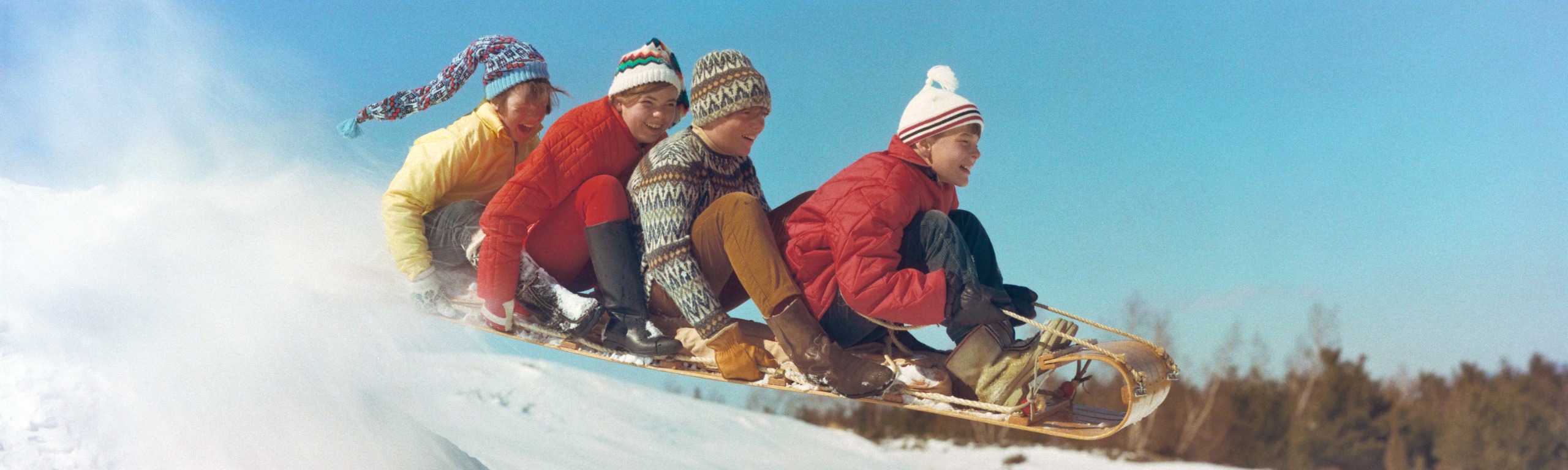 Four children in bright knitted bobble hats and quilted jackets, one red, one yellow, sit on a wooden toboggan as it flies over a snowy ridge against a clear blue sky, laughing