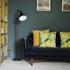 A dark green-painted living room with a dark blue velvet sofa with plump yellow cushions