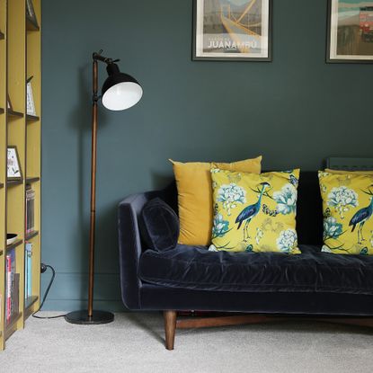 A dark green-painted living room with a dark blue velvet sofa with plump yellow cushions